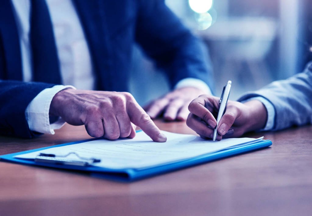 A man is pointing to a line on a contract that is being signed by someone else also sitting at the table