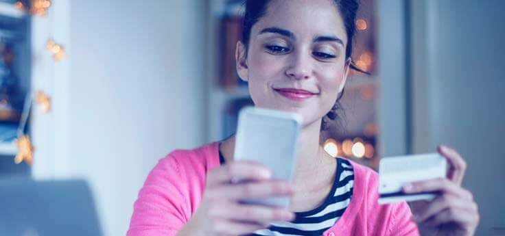 Lady in a pink cardigan looking at her smartphone in one hand and her credit card in the other