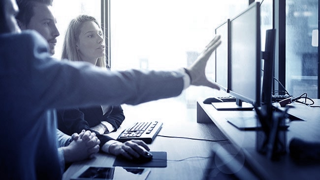 A group of three are gathered at a coworkers desk discussing the current project or assignment present on screen.