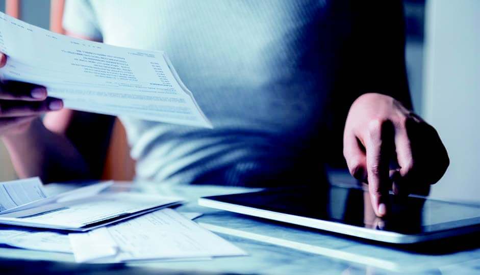 A woman holds a paper and is scrolling through her tablet at her desk.