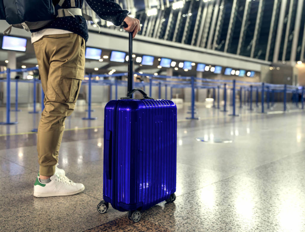 A man stands with his blue rolling suitcase in an airport, it is a shot from the waste down