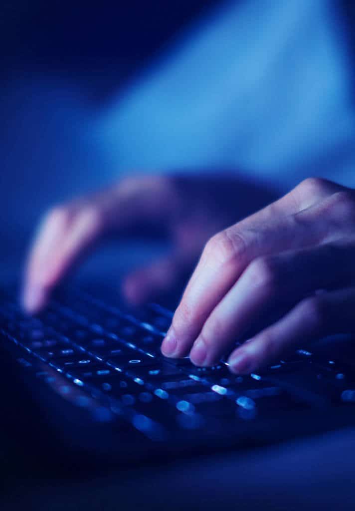 A close up shot of a person typing on their keyboard, the blue light of the computer screen lighting up their hands.