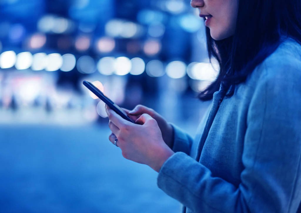 Person on the side of the street typing on their phone with a night city street is blurred out behind her